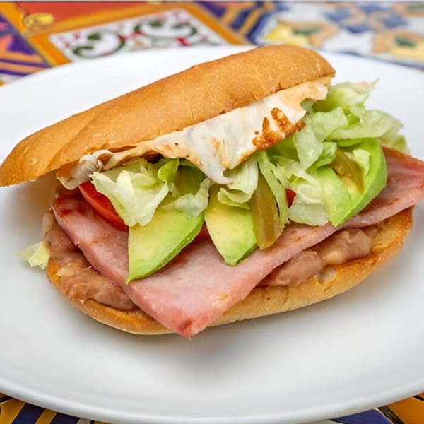 José's Torta Mexicana with ham, avocado, lettuce, tomato, beans, and melted cheese on a white plate.