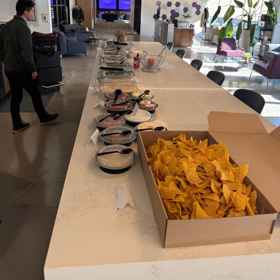 A long table filled with José's Torta Mexicana catering, including chips and dips, in a modern office.