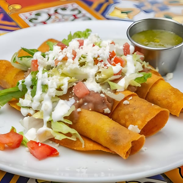 Three golden-fried taquitos topped with lettuce, sour cream, crumbled cheese, and diced tomatoes, with green salsa.